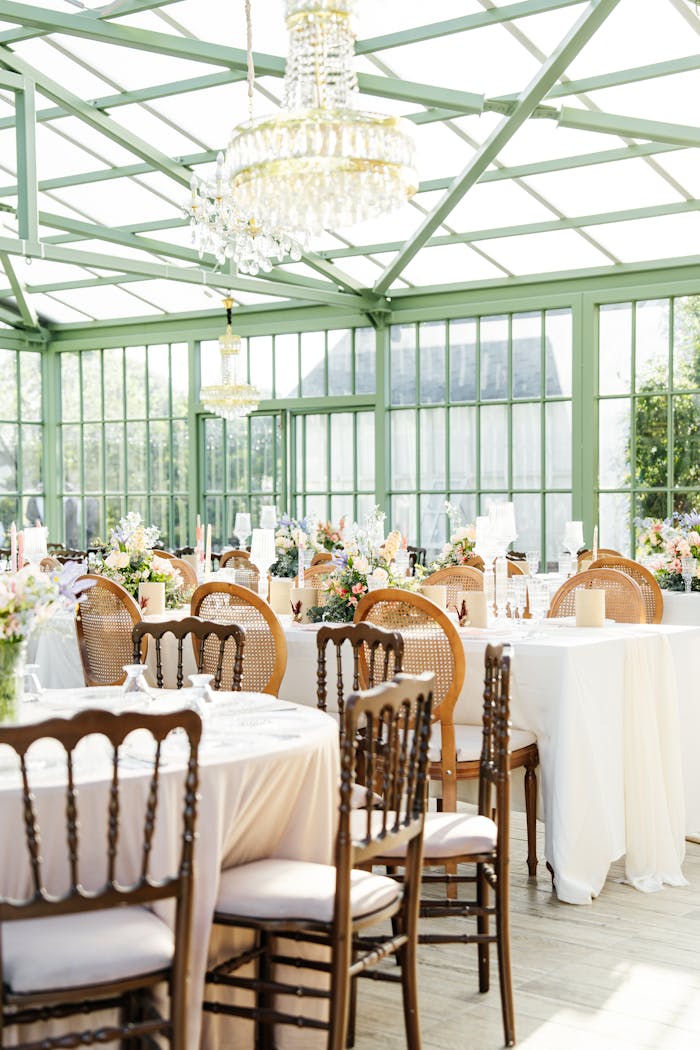 services-03 Sunlit glass venue set for an elegant wedding reception with floral decorations and chandeliers.